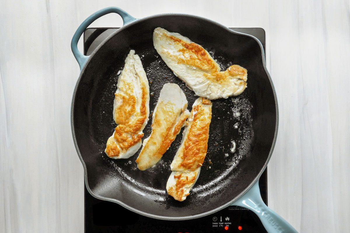 overhead shot of a black cast iron skillet with four chicken breasts cooking on a black induction cooktop;
