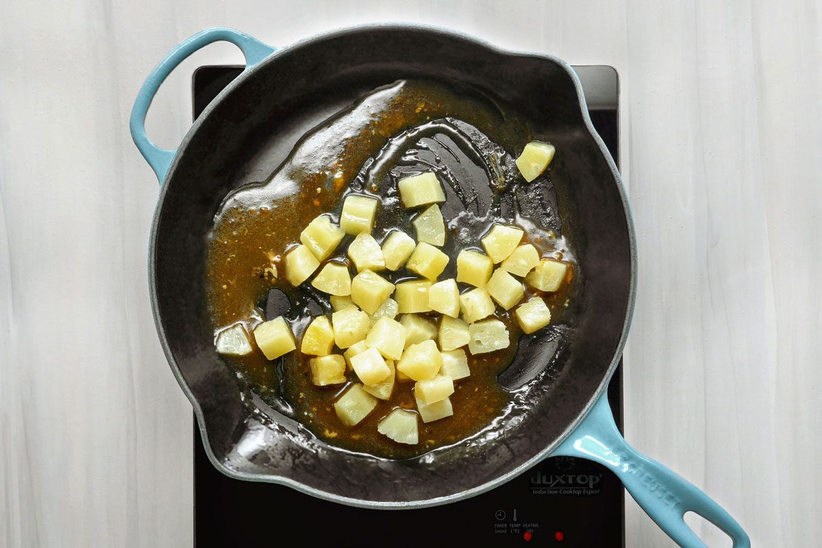 overhead shot of a cast iron skillet sitting on an induction cooktop; the skillet has a blue handle and is filled with cubed pineapple in a brown sauce; the skillet is on a black induction cooktop with a silver control panel
