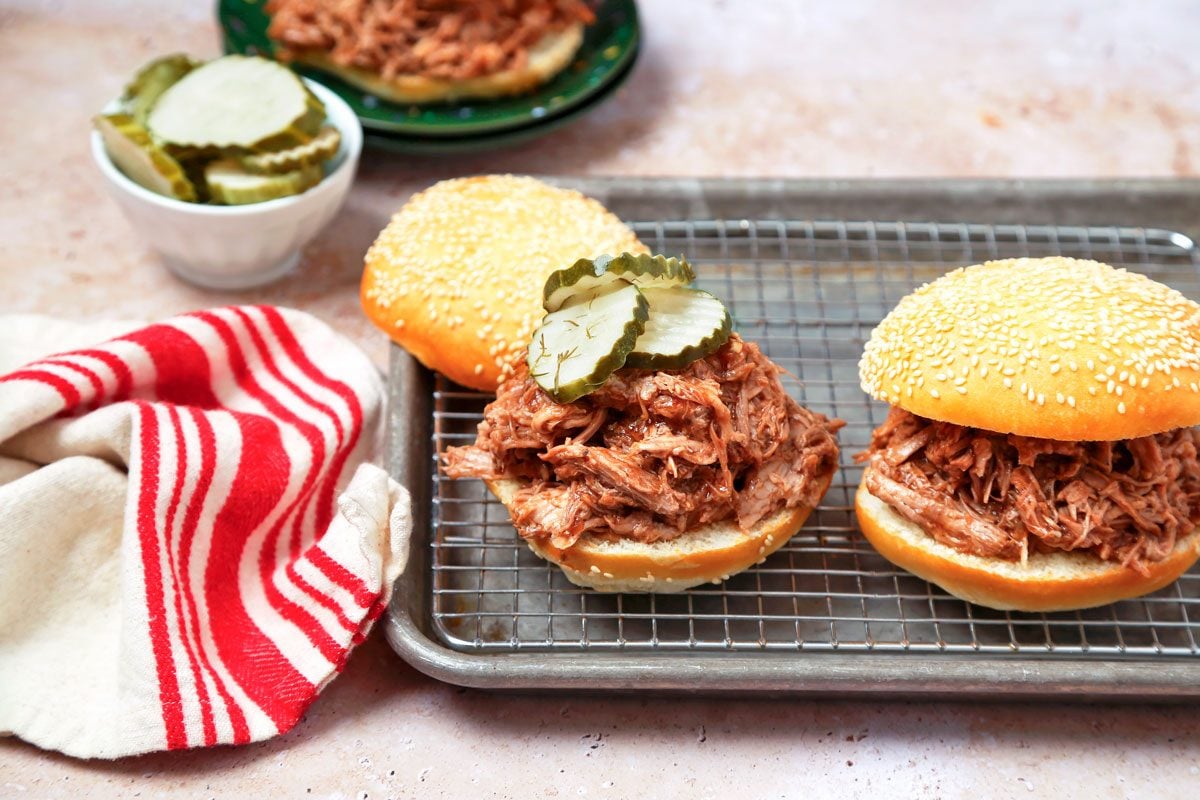 Pulled pork assembled into sandwiches on sesame seed buns with pickles.