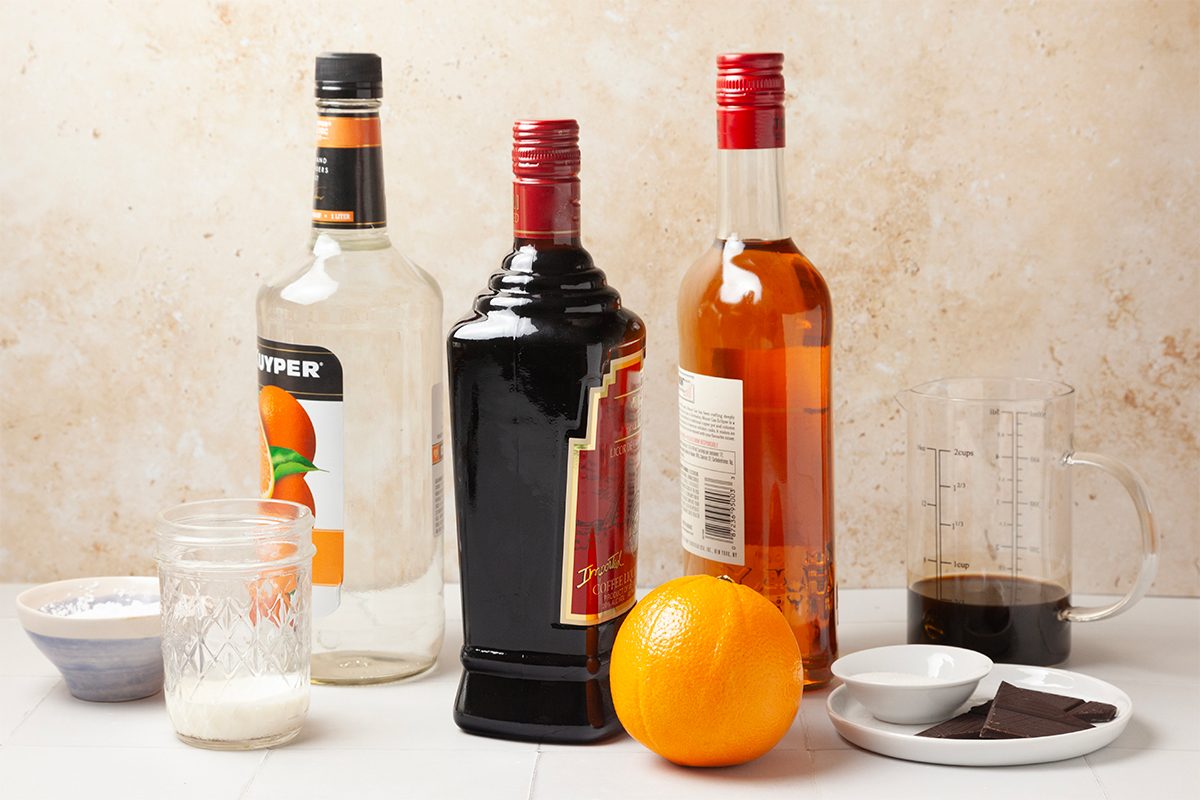 Three liquor bottles, an orange, a small bowl with white powder, a glass with dark liquid, and a white ramekin with two squares of dark chocolate are arranged on a countertop.