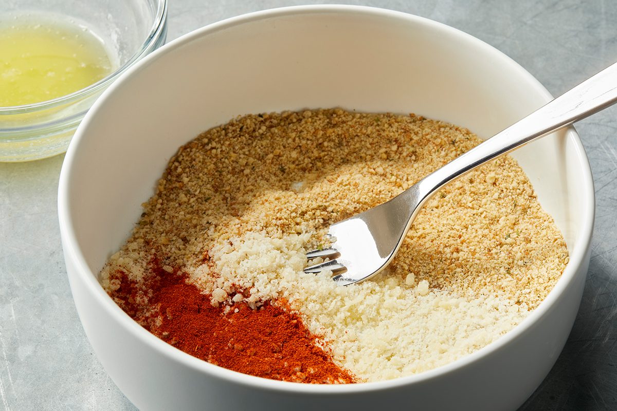 A white bowl contains a mix of breadcrumbs, grated parmesan cheese, and paprika, with a fork placed inside. A small bowl of egg white is seen in the background on a gray surface.