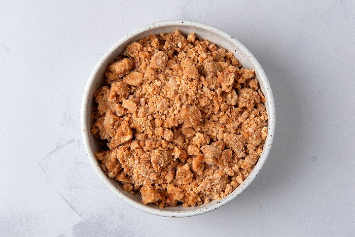 A bowl filled with crumbled brownish topping or mixture, possibly resembling streusel or a crumbly dessert component, placed on a light gray surface.