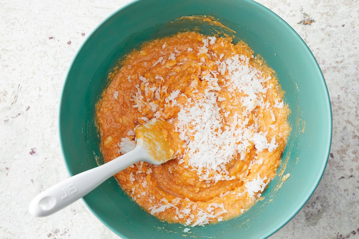 A teal bowl filled with an orange mixture, topped with grated white coconut. A white spoon rests in the mixture on the left side. The background is a light, textured surface.