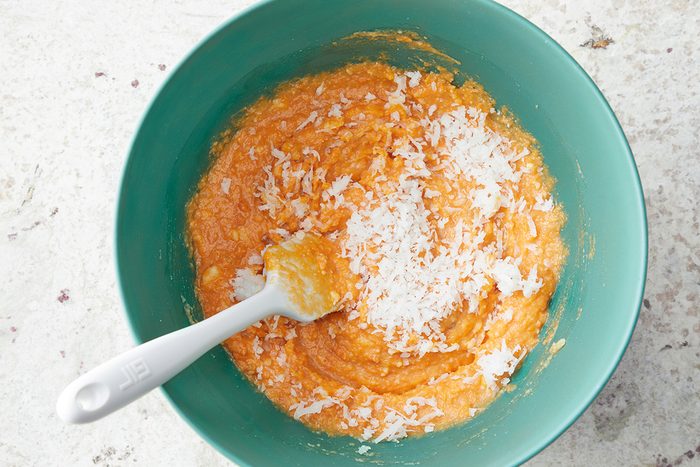 A teal bowl filled with an orange mixture, topped with grated white coconut. A white spoon rests in the mixture on the left side. The background is a light, textured surface.