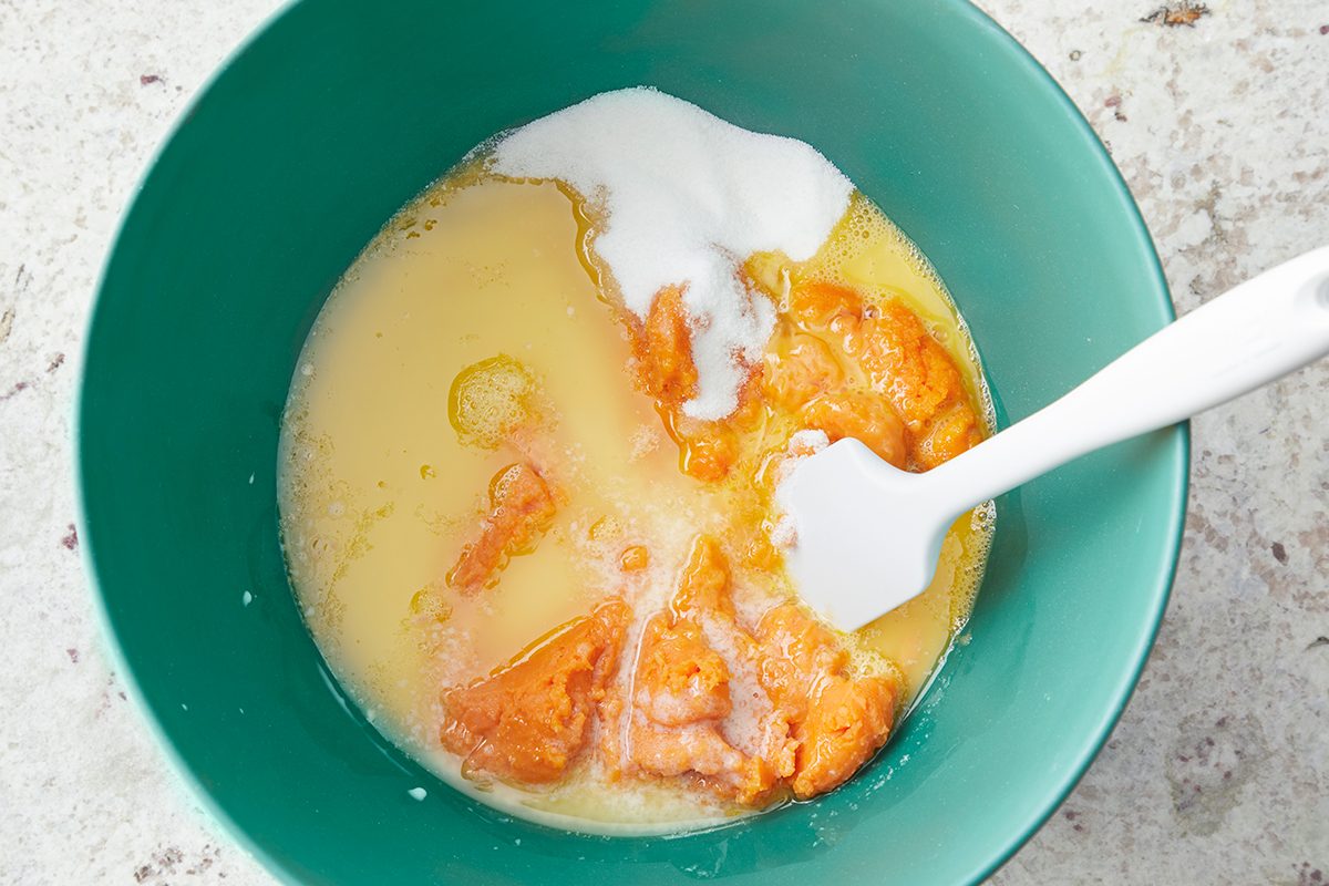 A green mixing bowl containing orange purée, sugar, and liquid ingredients, with a white spatula on the right side, resting inside. The mixture is unblended, showing distinct ingredients.