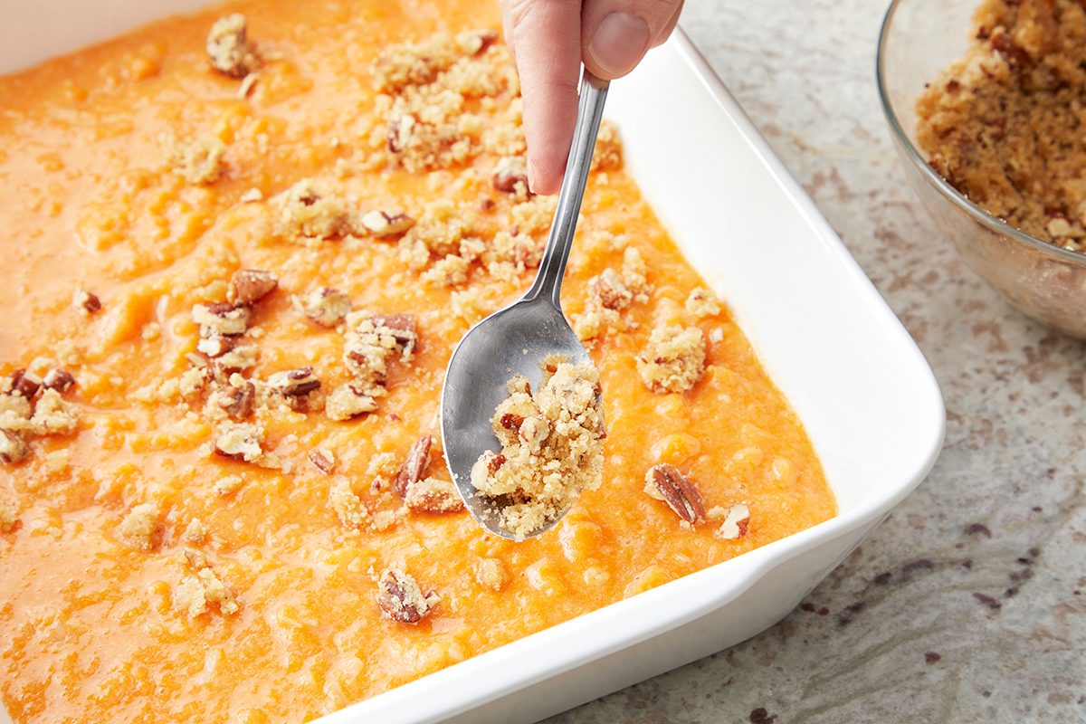 A hand holds a spoon, sprinkling a crumbly mixture with pecans over a creamy, orange sweet potato casserole in a white rectangular dish. A small bowl with more topping is nearby on a stone surface.