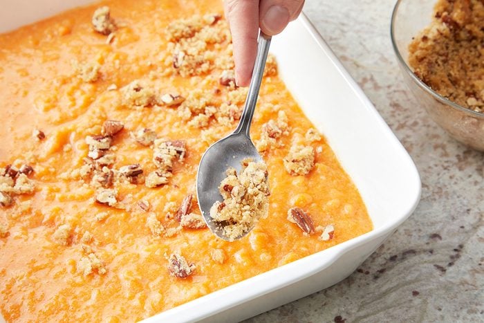 A hand holds a spoon, sprinkling a crumbly mixture with pecans over a creamy, orange sweet potato casserole in a white rectangular dish. A small bowl with more topping is nearby on a stone surface.