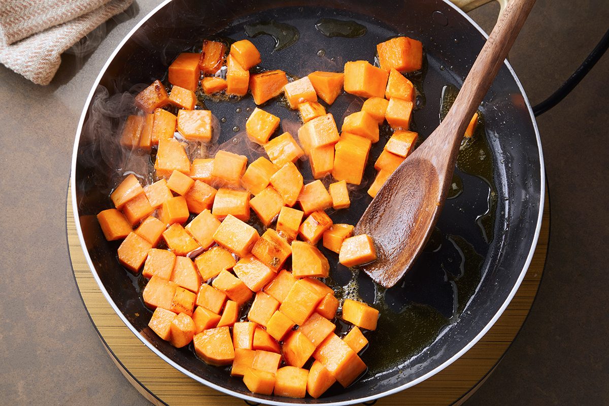 Cubed sweet potatoes cooking in a skillet with olive oil, being stirred by a wooden spoon. The cubes are partially browned, and steam is visible rising from the pan. The skillet sits on a wooden surface.