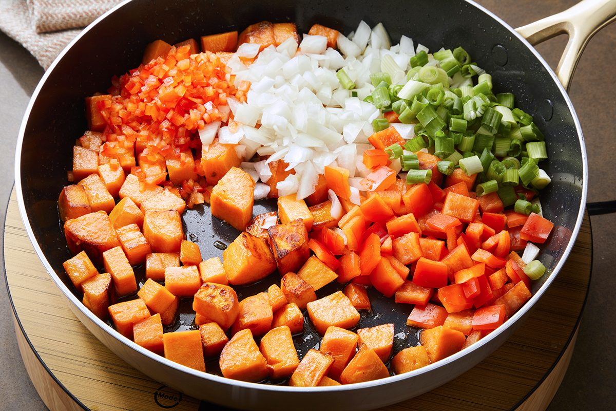 A skillet with chopped sweet potatoes, red bell pepper, onion, and celery. The vegetables are arranged separately in sections within the pan, ready for cooking.