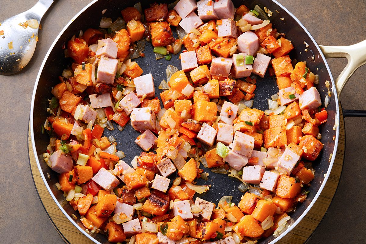 A skillet filled with a cooked mixture of diced ham, sweet potatoes, onions, and green peppers. A spoon rests on the surface next to the skillet. The ingredients appear seasoned and sautéed.