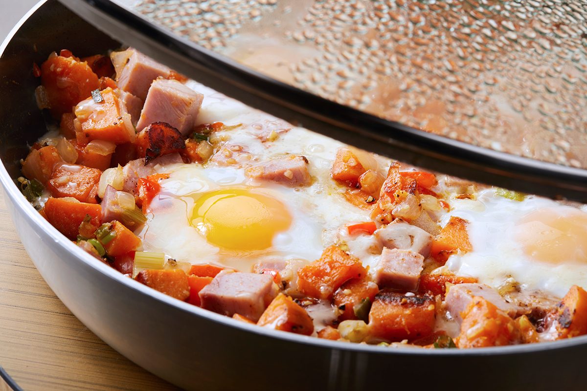 A skillet filled with sweet potato hash, featuring visible cubes of sweet potato, chunks of ham, diced vegetables, and two sunny-side-up eggs. A glass lid partially covers the pan, with condensation on the surface.