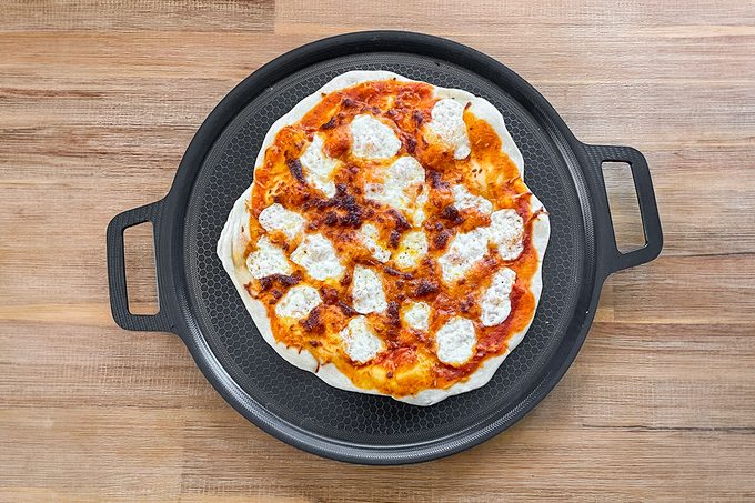 A homemade pizza with a golden crust, topped with melted cheese and tomato sauce, on a round pizza pan. The pan has two handles and is placed on a wooden table.