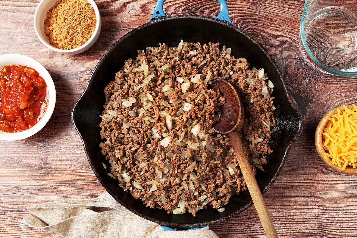 overhead shot of a cast iron skillet filled with cooked ground beef and onions, sitting on a wooden table; a wooden spoon is in the skillet and a bowl of shredded cheese is to the right of the skillet; a small bowl of salsa and a bowl of ground spices is to the left of the skillet; a glass measuring cup filled with liquid is to the right of the skillet, on the table; there is a light tan cloth napkin folded over the left side of the skillet