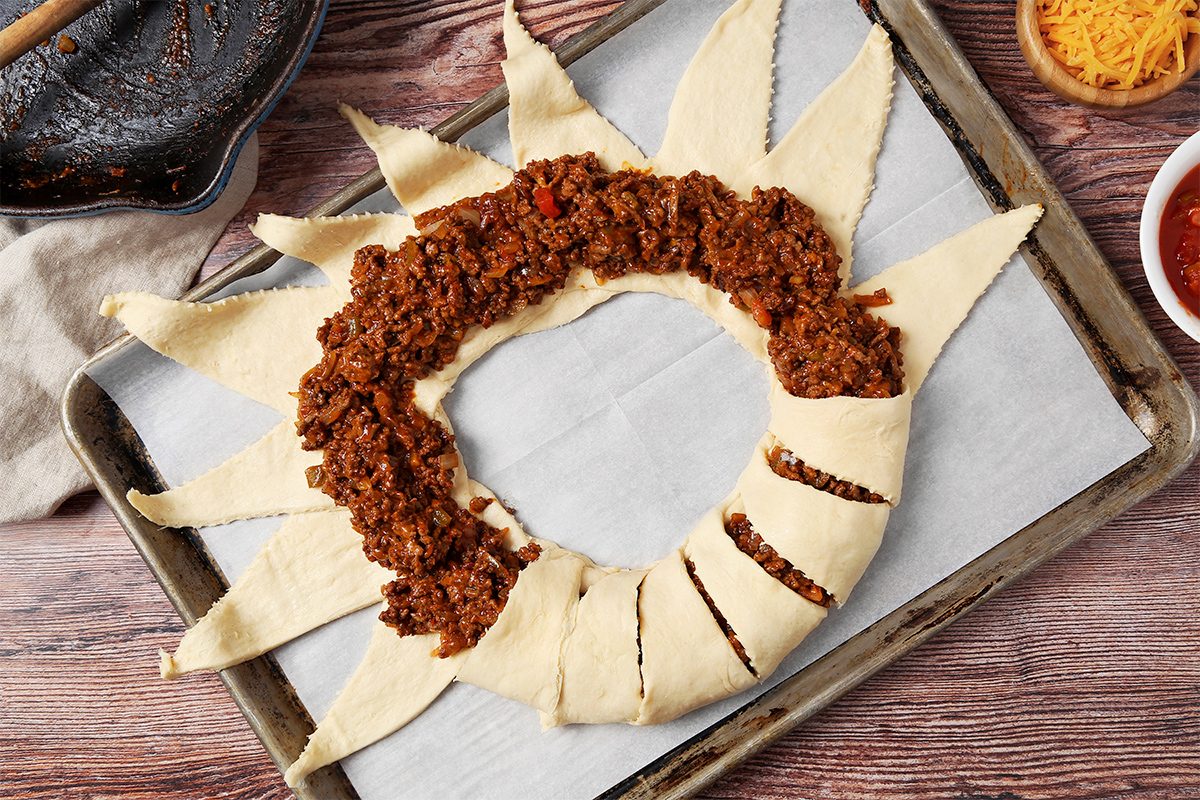 overhead shot of a baking sheet with a crescent roll dough shaped into a ring and filled with a meat sauce; the baking sheet is on a wooden table and has parchment paper on it, the crescent roll dough is shaped into a ring and filled with a meat sauce; on the left side of the image, a cast iron skillet is shown and on the right, there are two bowls - one with grated cheese and one with salsa