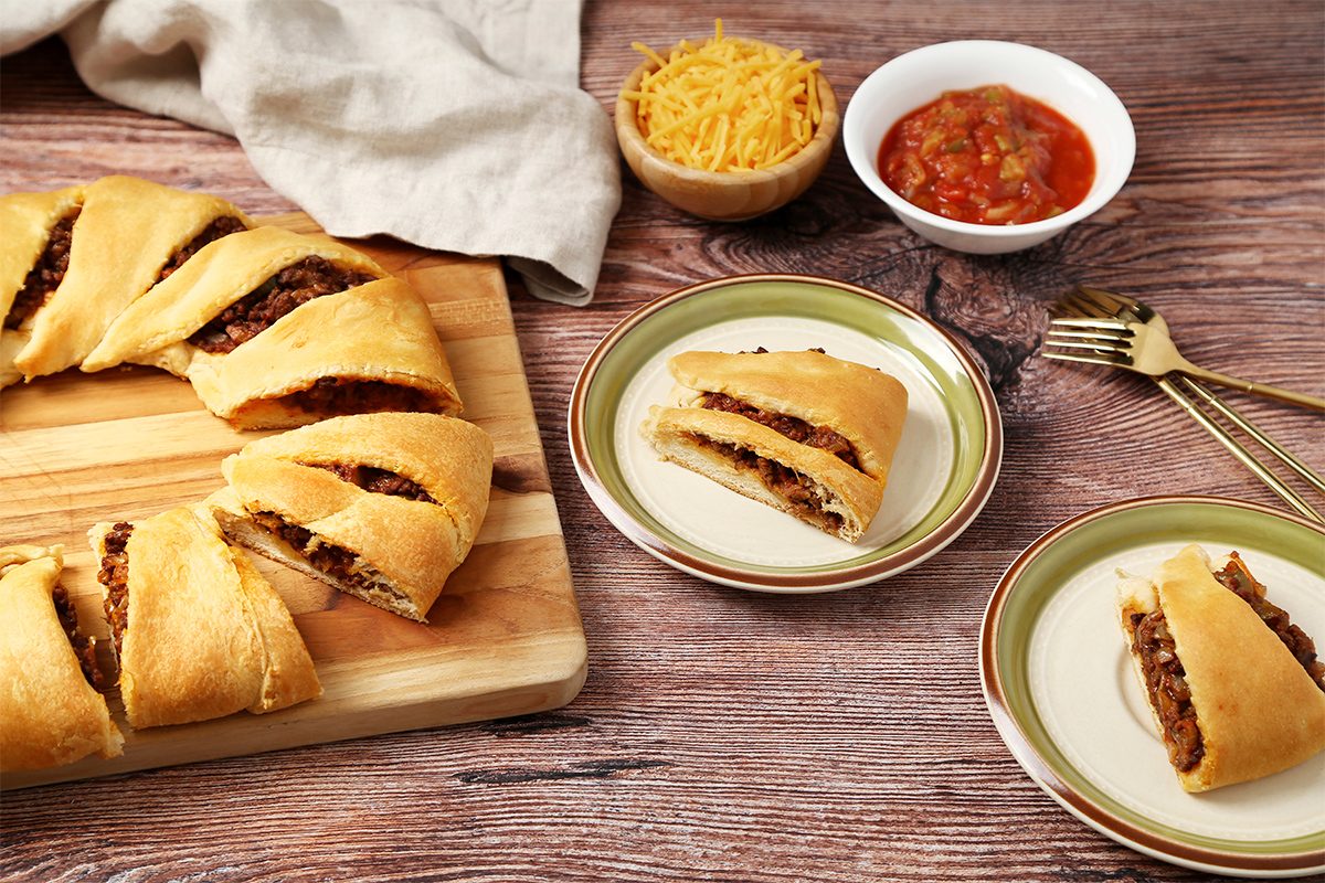 3/4th shot of a wooden cutting board is in the center with a golden brown pastry ring with filling inside, a portion of it is separated into 8 triangular pieces; a white ceramic bowl with red salsa, a bowl with grated cheese, and two green and brown patterned plates, each with a single piece of the pastry ring; golden forks are lying on the side; the background is a wooden surface with a light brown linen cloth to the left