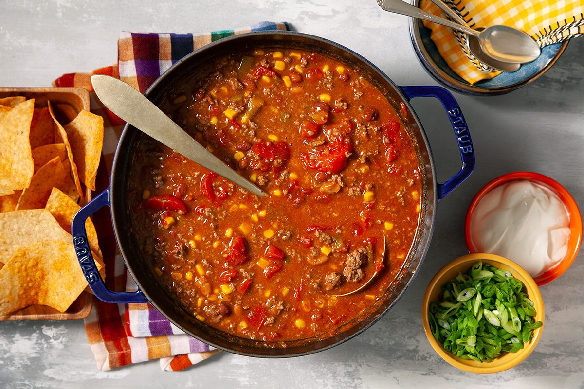 A pot of chili with tomatoes, corn, and beans, served with a ladle. Tortilla chips are on the side, and small bowls of chopped green onions and sour cream are ready for garnishing. A checkered napkin and spoons complete the setting.