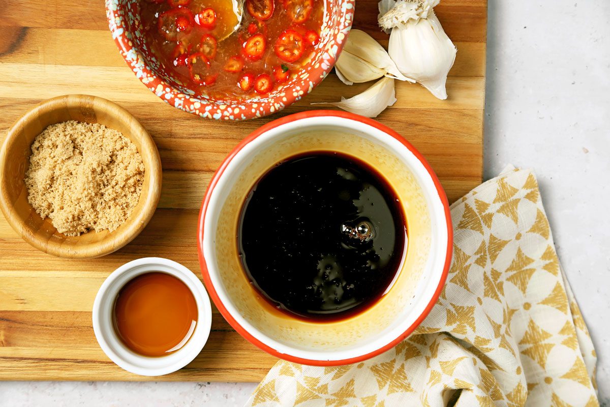 A wooden board holds a bowl of brown sugar, a bowl of soy sauce, a small dish of honey, and a clay bowl of chili sauce with garlic cloves beside it. A cream-colored cloth with a star pattern is on the side.