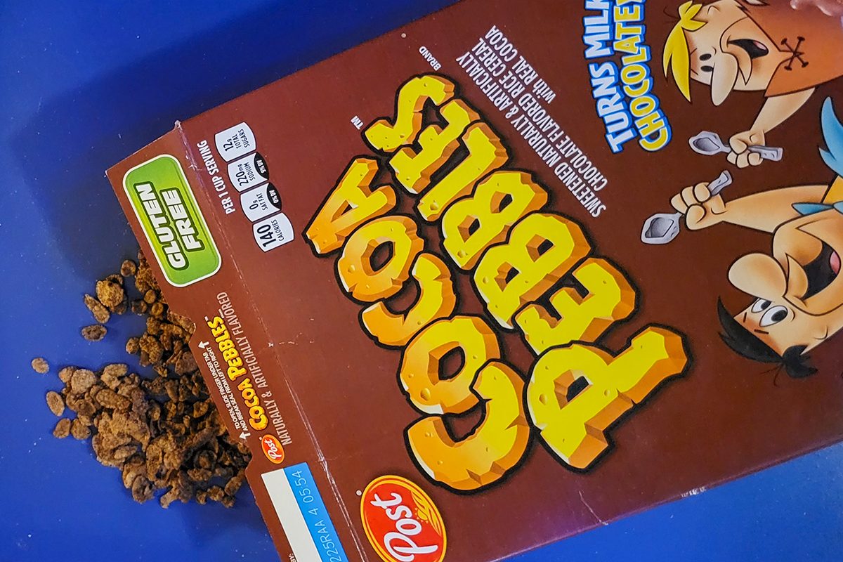 A box of Cocoa Pebbles cereal, featuring colorful branding and cartoon characters, lies on its side with some cereal pieces spilled next to it on a blue surface.