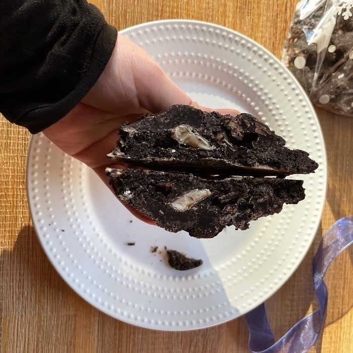 Hand holds a cross section of a dark chocolate cookie with toffee chips above a white plate and wooden table