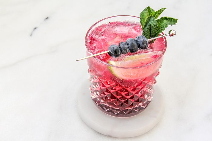 A refreshing pink cocktail with ice, garnished with blueberries on a skewer and a sprig of mint. The drink is served in a textured glass on a marble surface.