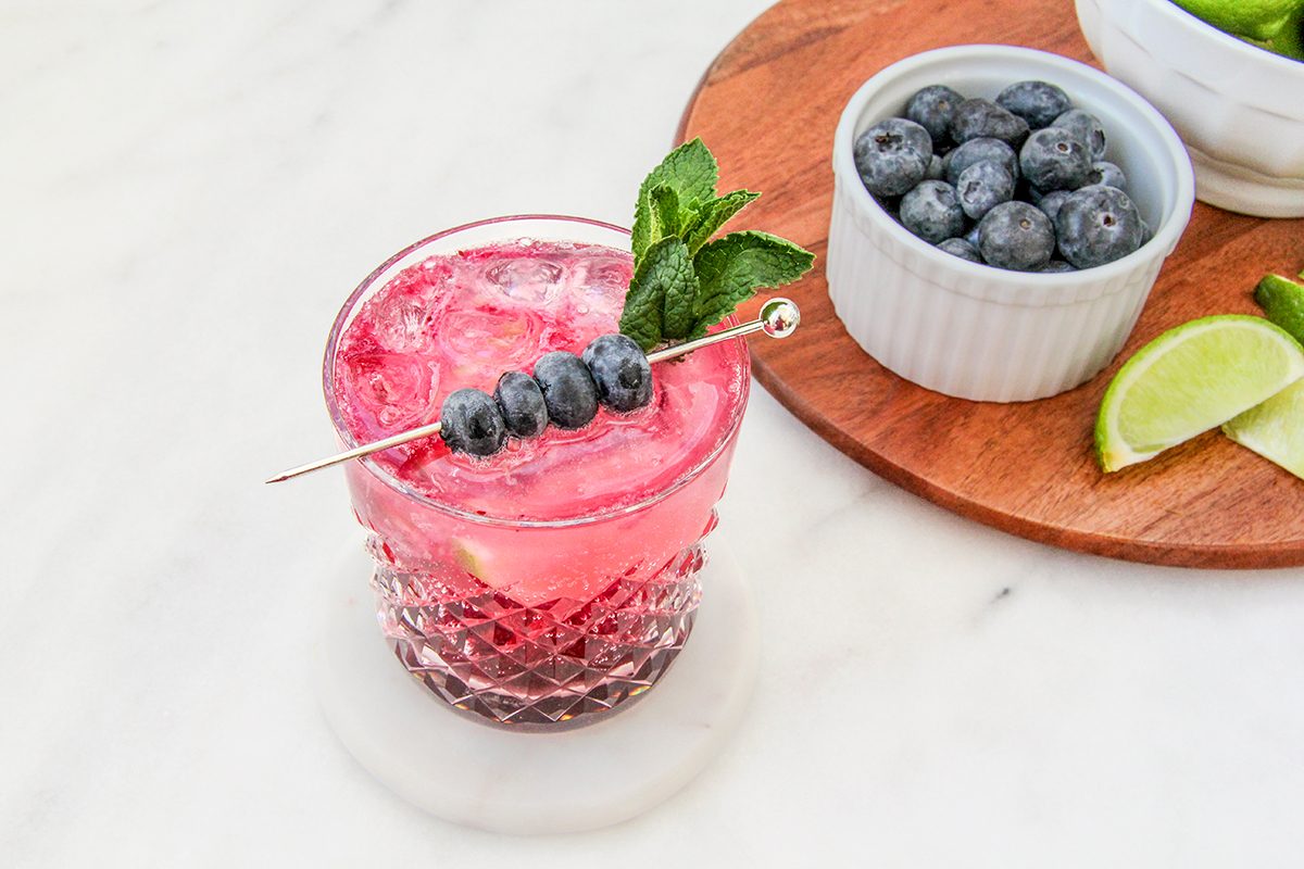 A glass of pink cocktail with mint leaves and blueberries on a skewer. It sits on a white coaster. Nearby, a wooden board holds a dish of blueberries, lime wedges, and a bowl. The background is a light-colored surface.