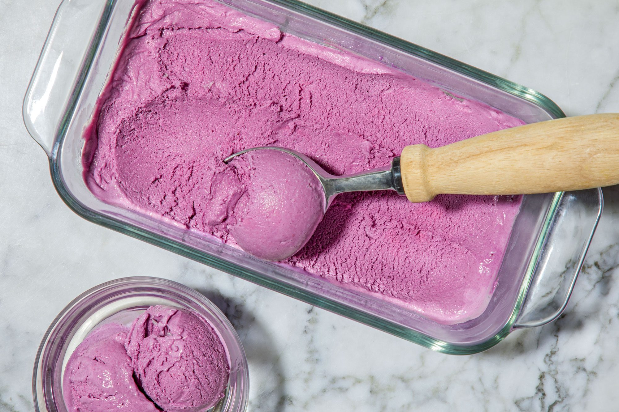 overhead horizontal shot of Blueberry Ice Cream in a clear glass container; A metallic ice cream scoop with a wooden handle is positioned on the side, having just carved out a round scoop of the ice cream; Nearby, a small dish holds additional scoops of the same ice cream