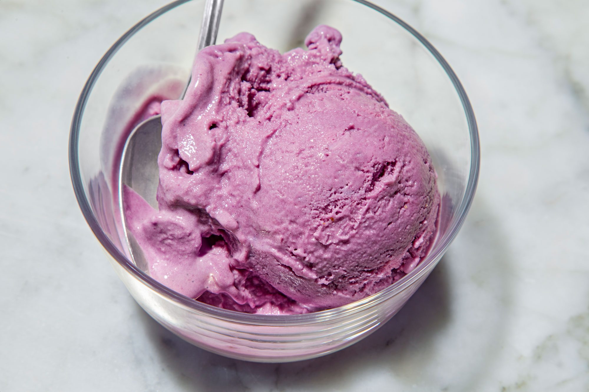 closeup shot of Blueberry Ice Cream scoop in a clear glass bowl with a silver spoon resting inside the bowl, the bowl is placed over white marble background