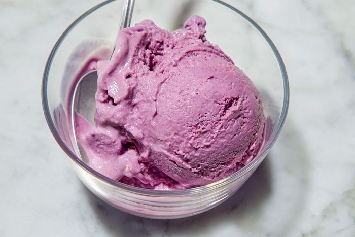 closeup shot of Blueberry Ice Cream scoop in a clear glass bowl with a silver spoon resting inside the bowl, the bowl is placed over white marble background