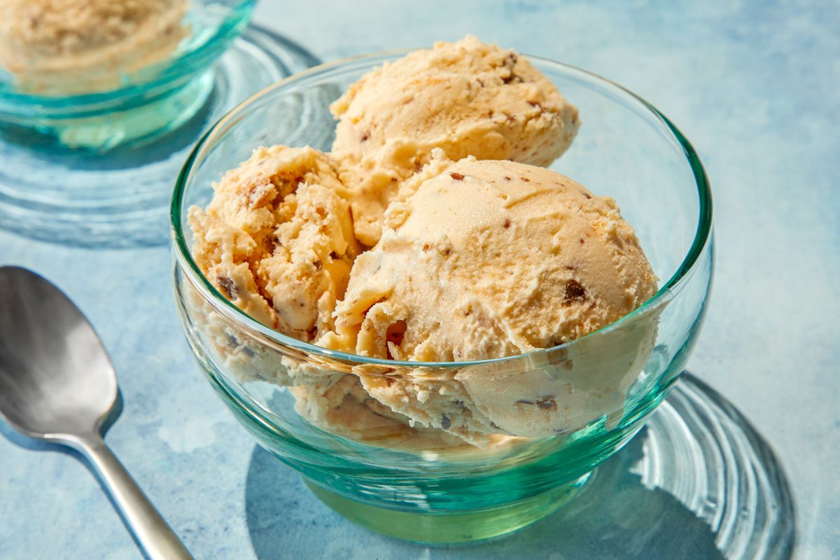 closeup shot of serving of Butterfinger Ice Cream in a clear glass bowl; in the background, another bowl of ice cream can be seen, along with a shiny metallic spoon resting beside the main bowl; the backdrop is a soft blue