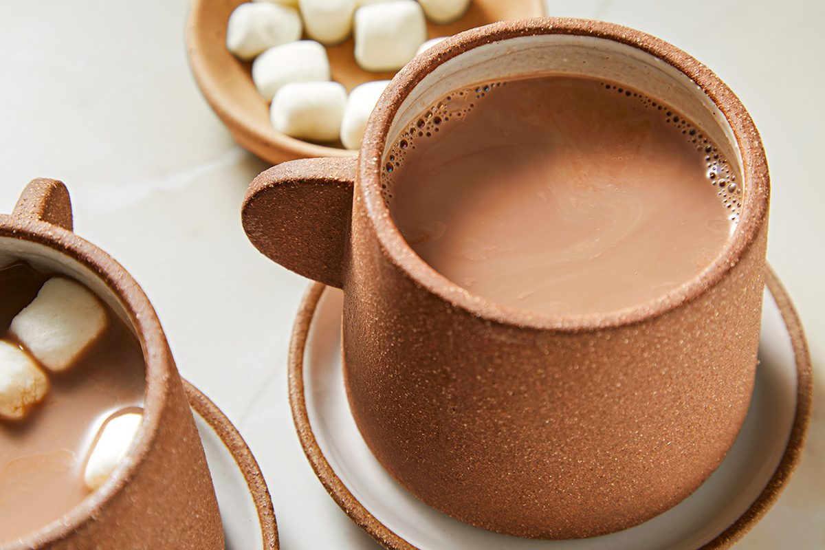 A ceramic mug filled with hot chocolate sits on a saucer. Nearby, a bowl of marshmallows and part of another mug with marshmallows inside can be seen. The scene suggests a cozy and inviting setting for enjoying a warm drink.