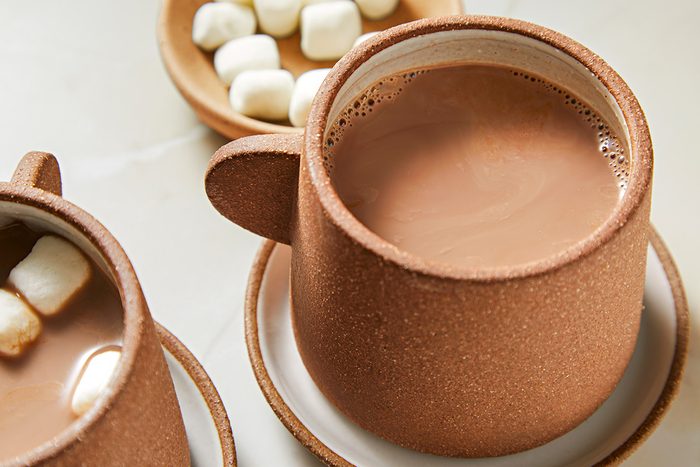 A ceramic mug filled with hot chocolate sits on a saucer. Nearby, a bowl of marshmallows and part of another mug with marshmallows inside can be seen. The scene suggests a cozy and inviting setting for enjoying a warm drink.