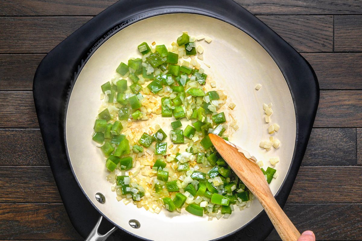 overhead shot of diced green peppers, onions, and minced garlic being sauteed in a white pan