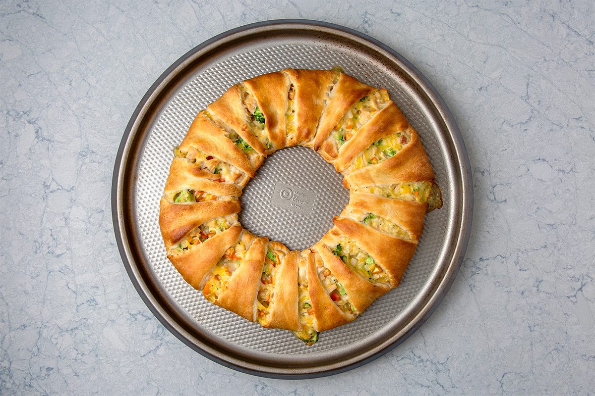 A round baking tray holds a golden-brown crescent roll ring with visible fillings of chicken, melted cheese, and chopped green onions. The ring is evenly baked, showing a flaky texture against a light gray background.