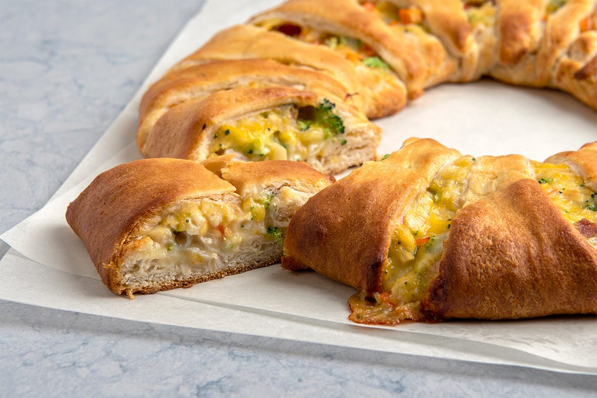 A golden-brown crescent ring with visible filling of chicken, cheese, and vegetables sits on parchment paper. One slice is cut, showing the savory filling. The background surface is light and speckled.