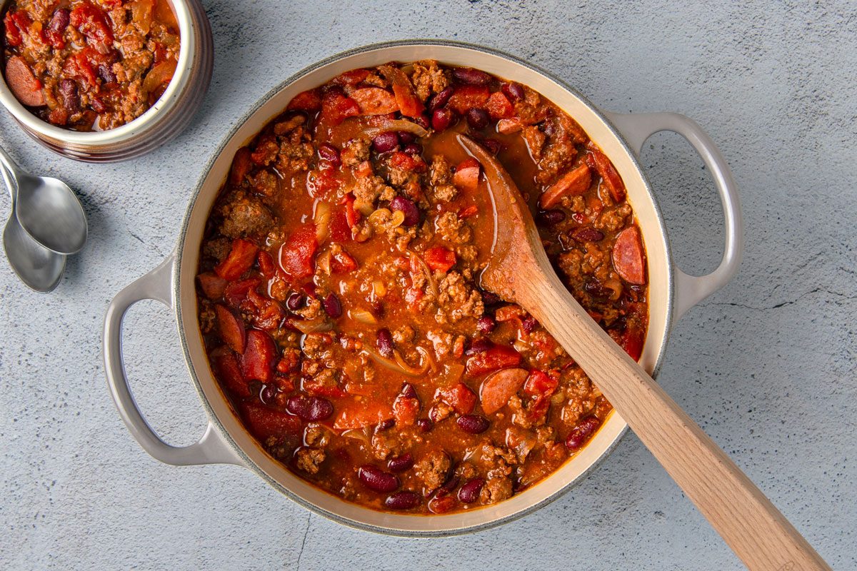 overhead shot of a pot of chili; the pot is white with a light grey handle and a wooden spoon is resting on top of the chili, there is a small brown pot of chili in the top left corner of the image, and two spoons are resting near it