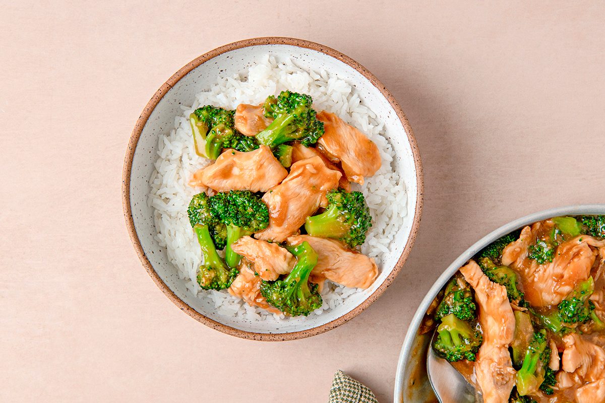 A bowl of rice topped with cooked chicken pieces and broccoli, served on a beige surface. Part of another dish with the same ingredients is partially visible next to it.
