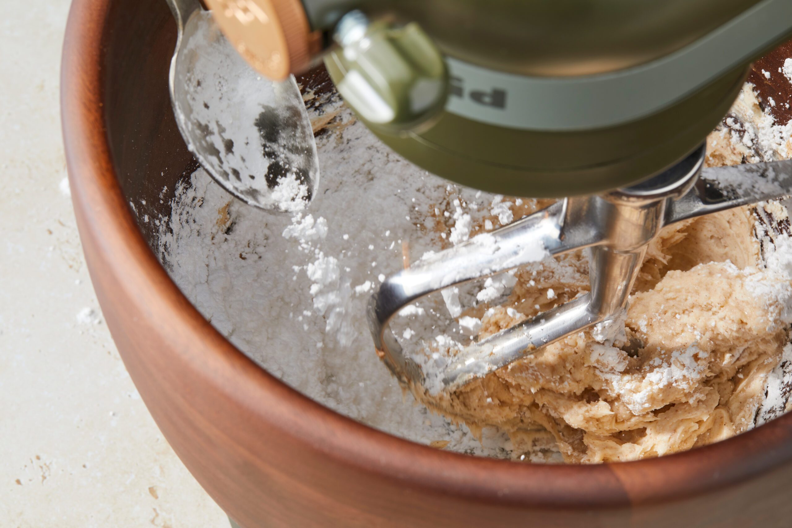 A top-down view of an electric mixer blending cookie dough in a wooden bowl. The metal beaters are coated with dough, and flour is sprinkled on the sides of the bowl.