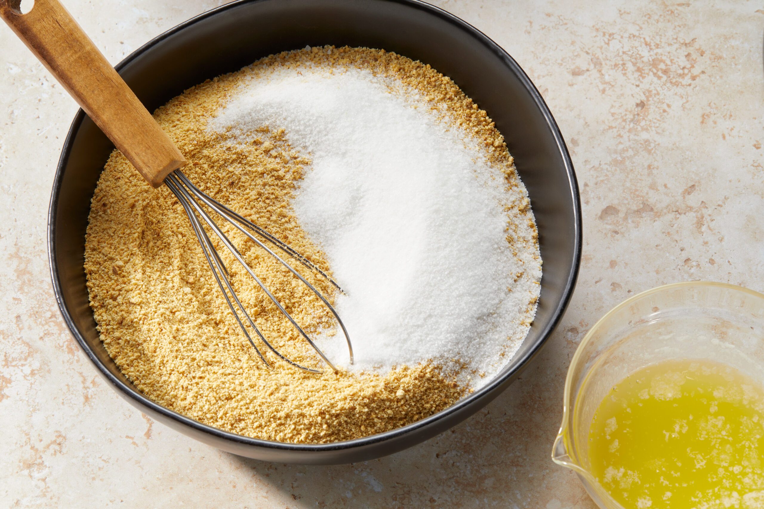 A black bowl with graham cracker crumbs and sugar is partially mixed with a whisk. Next to the bowl is a small glass container holding melted butter. The items are placed on a light stone surface.