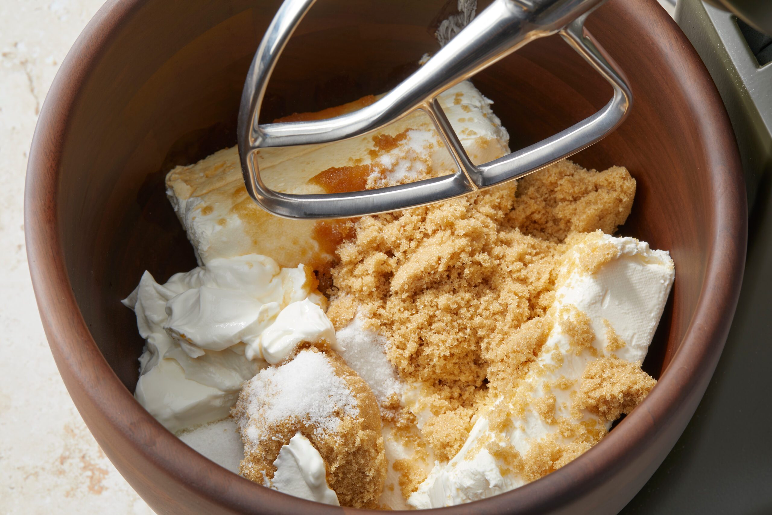 A mixing bowl contains blocks of cream cheese, brown sugar, a cream-like ingredient, and a hint of vanilla extract. A metal mixer attachment is positioned above the ingredients, ready for use.
