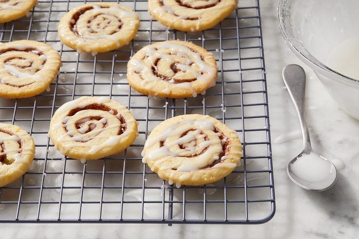 Cinnamon roll cookies with icing sit on a cooling rack. A glass bowl with a spoon beside it rests on a marble countertop.