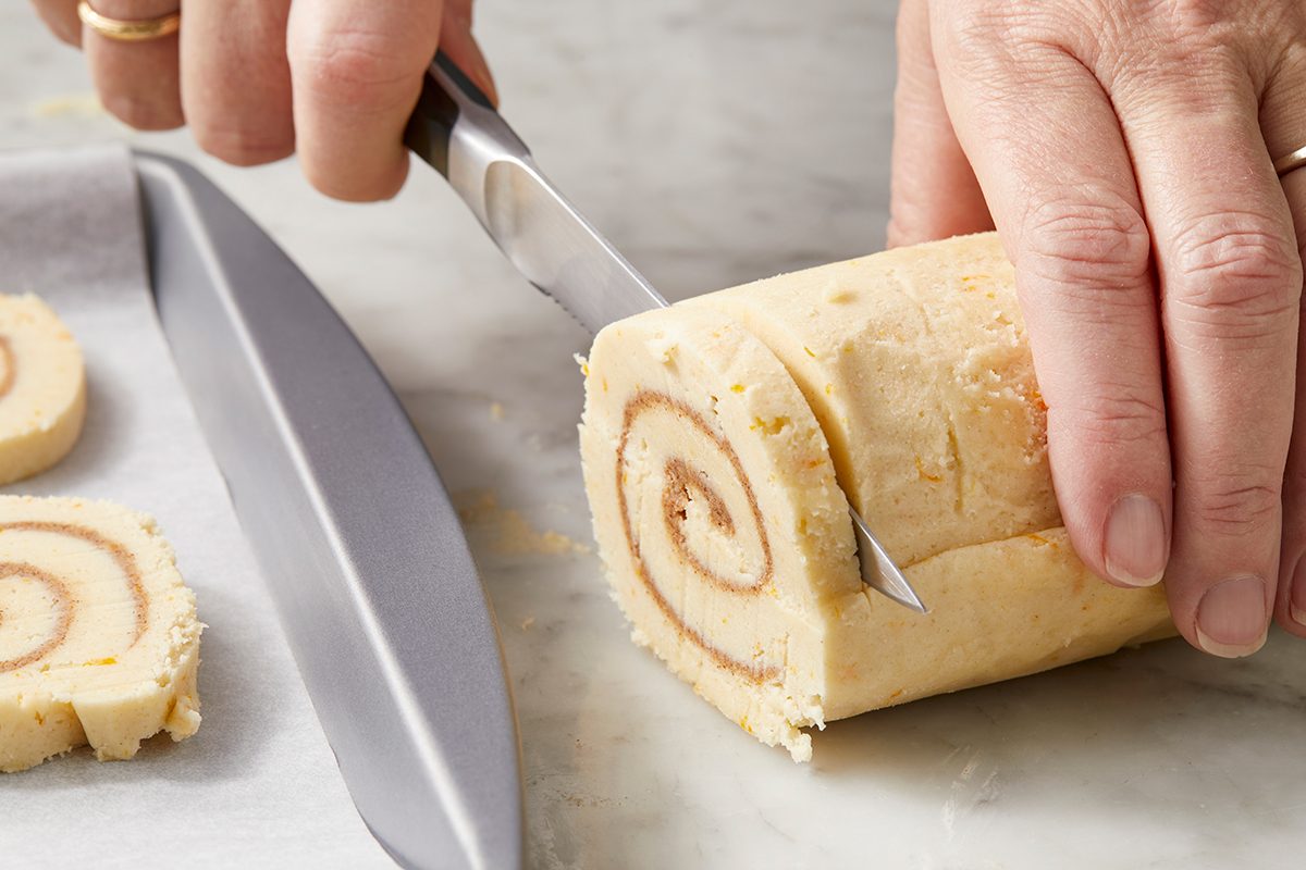 Close-up of a person slicing a roll of uncooked cinnamon dough on a marble surface. The roll has visible spirals, and a sharp knife is cutting through, with a sheet of parchment paper nearby.