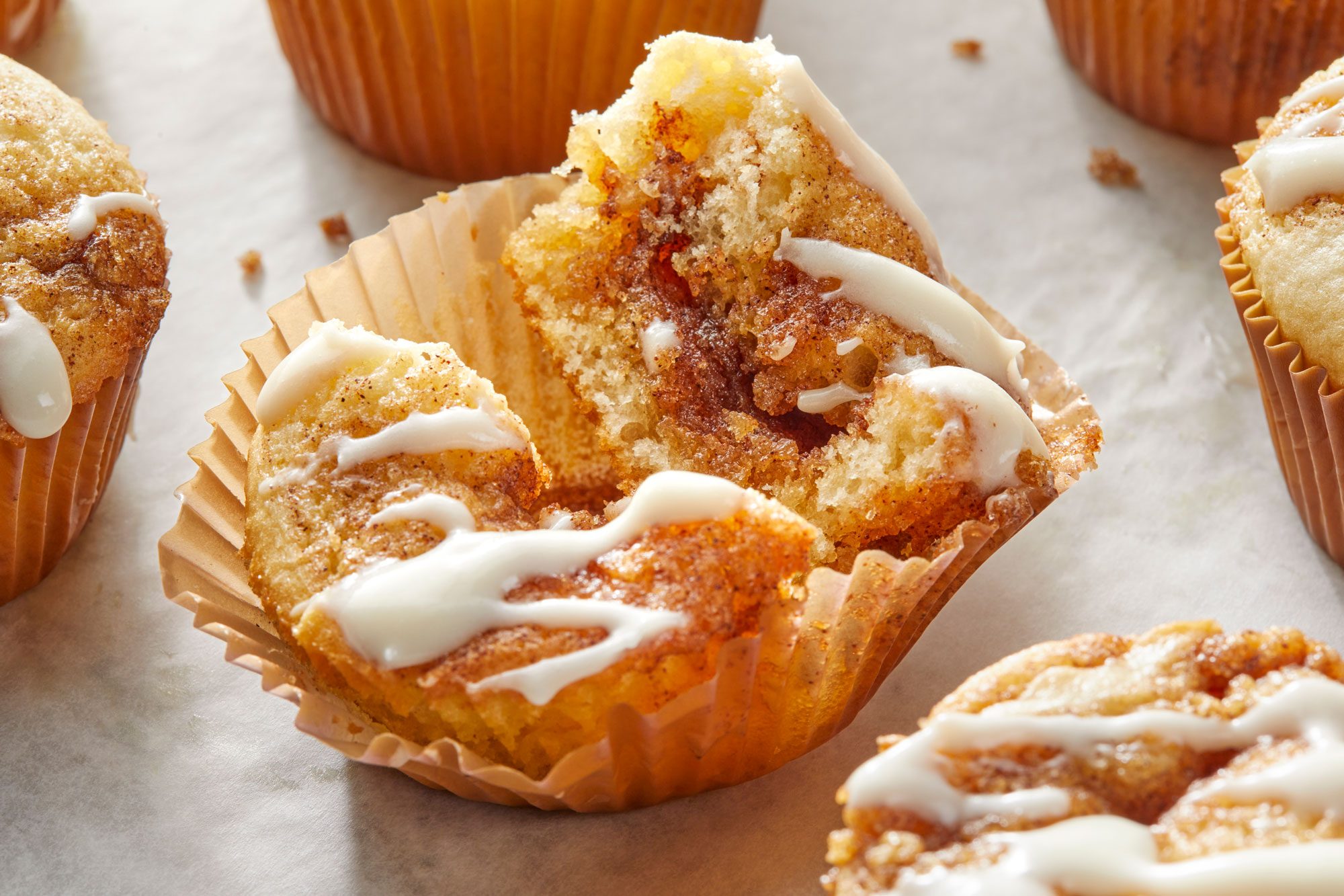 a closeup shot of a partially unwrapped baked good on a light colored surface; the baked good, likely a muffin or cupcake, is presented in a brown paper liner; is drizzled with a white icing in a flowing, the top of the muffin has been broken or torn open, revealing a cinnamon swirl pattern inside, the surface on which the pastry rests is a light gray