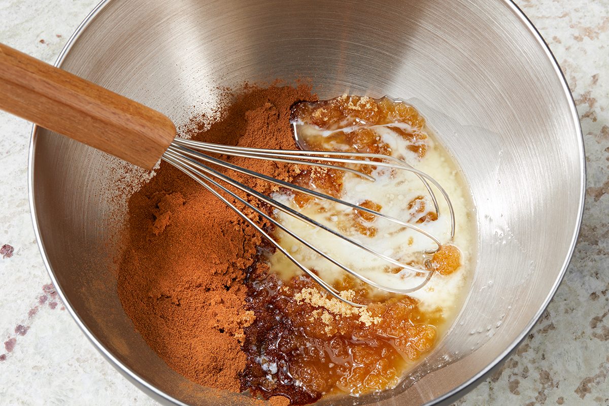 A metal mixing bowl contains a wooden-handled whisk, wet and dry ingredients including spices, brown sugar, and a sticky liquid. The bowl is set on a light-colored marbled countertop.