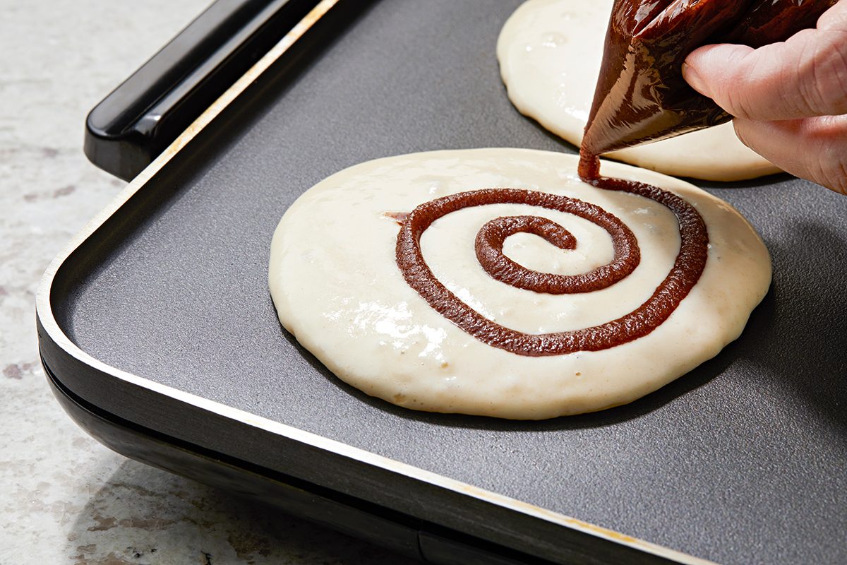 Close-up of pancake batter on a griddle. A hand is piping a swirl of chocolate batter onto the pancake. The griddle is on a gray countertop.