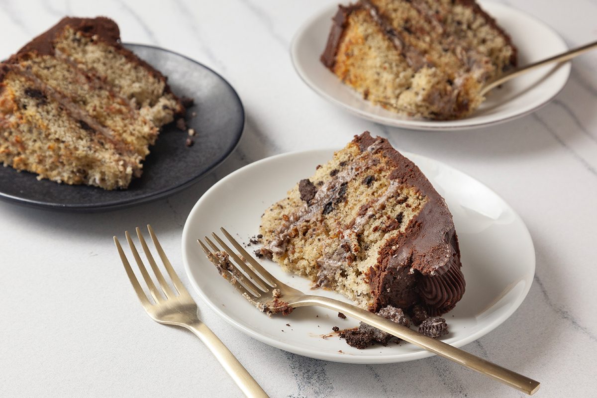 Slices of a layered chocolate cake with swirled frosting are placed on white and black plates. Each plate has a fork beside the cake, atop a white marble surface.
