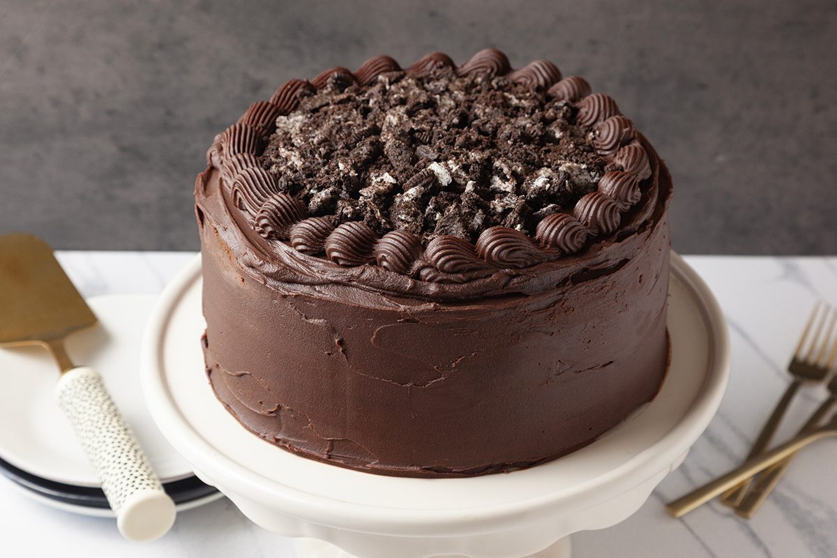 Chocolate cake with creamy frosting topped with crumbled cookies, displayed on a white cake stand. A gold cake server and forks are set beside the stand on a marbled surface.
