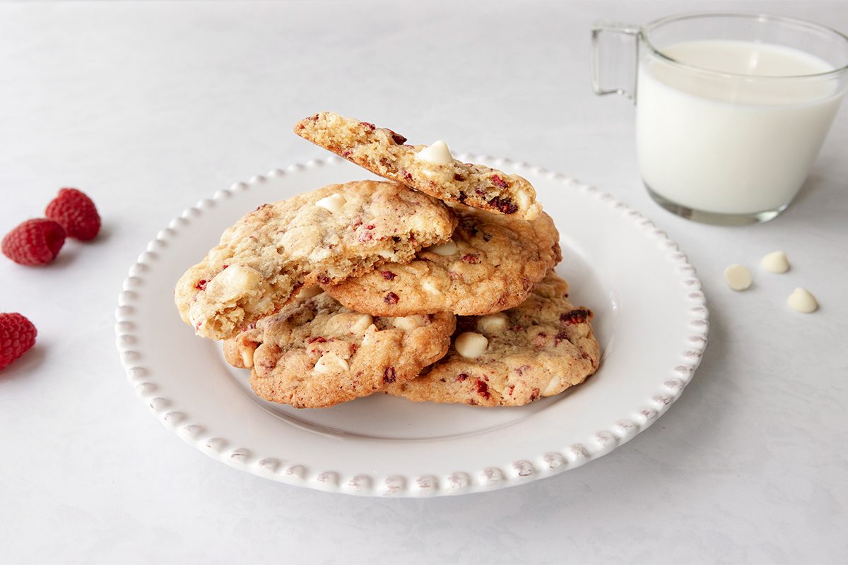 A plate of white chocolate raspberry cookies is stacked on a white plate with a beaded edge. Nearby are three raspberries and a glass cup filled with milk on a light gray surface.