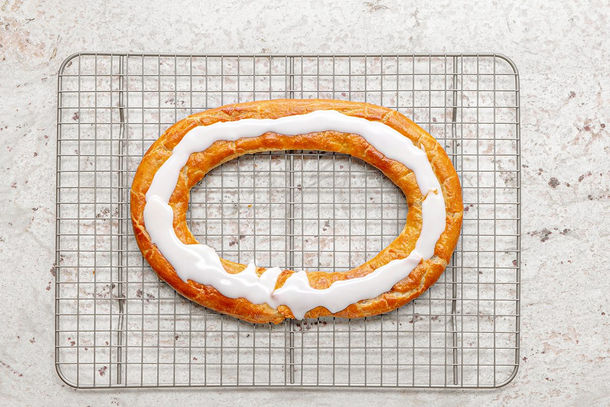 A freshly baked ring-shaped pastry with white icing drizzled on top sits on a cooling rack against a light, textured background.