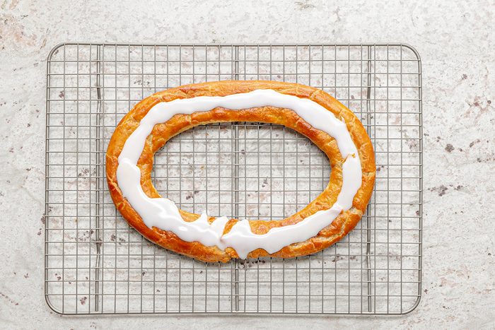 A freshly baked ring-shaped pastry with white icing drizzled on top sits on a cooling rack against a light, textured background.