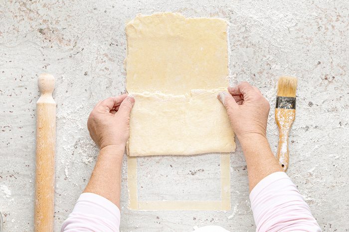 Hands folding a sheet of dough on a floured surface with a rolling pin and a pastry brush nearby.